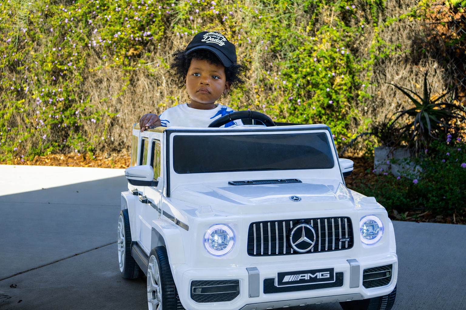 A child in a black Stunting Like My Daddy hat riding in a white toy SUV